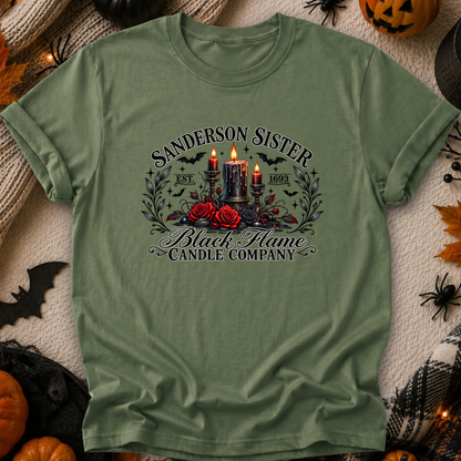An olive green graphic t-shirt laid flat on a cozy, Halloween-themed background. The shirt features a gothic-style graphic with the text 'SANDERSON SISTER' and 'Black Flame Candle Company' surrounding an illustration of three lit black candles, red roses, and bats.