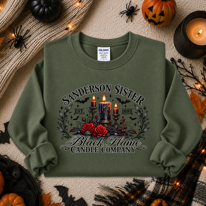 An olive green crewneck sweatshirt with a Halloween-themed graphic print reading 'Sanderson Sister Black Flame Candle Company' around an illustration of candles, roses, and bats. The sweatshirt is displayed in a flat-lay arrangement with seasonal props including pumpkins, a candle, and a cozy knit blanket.