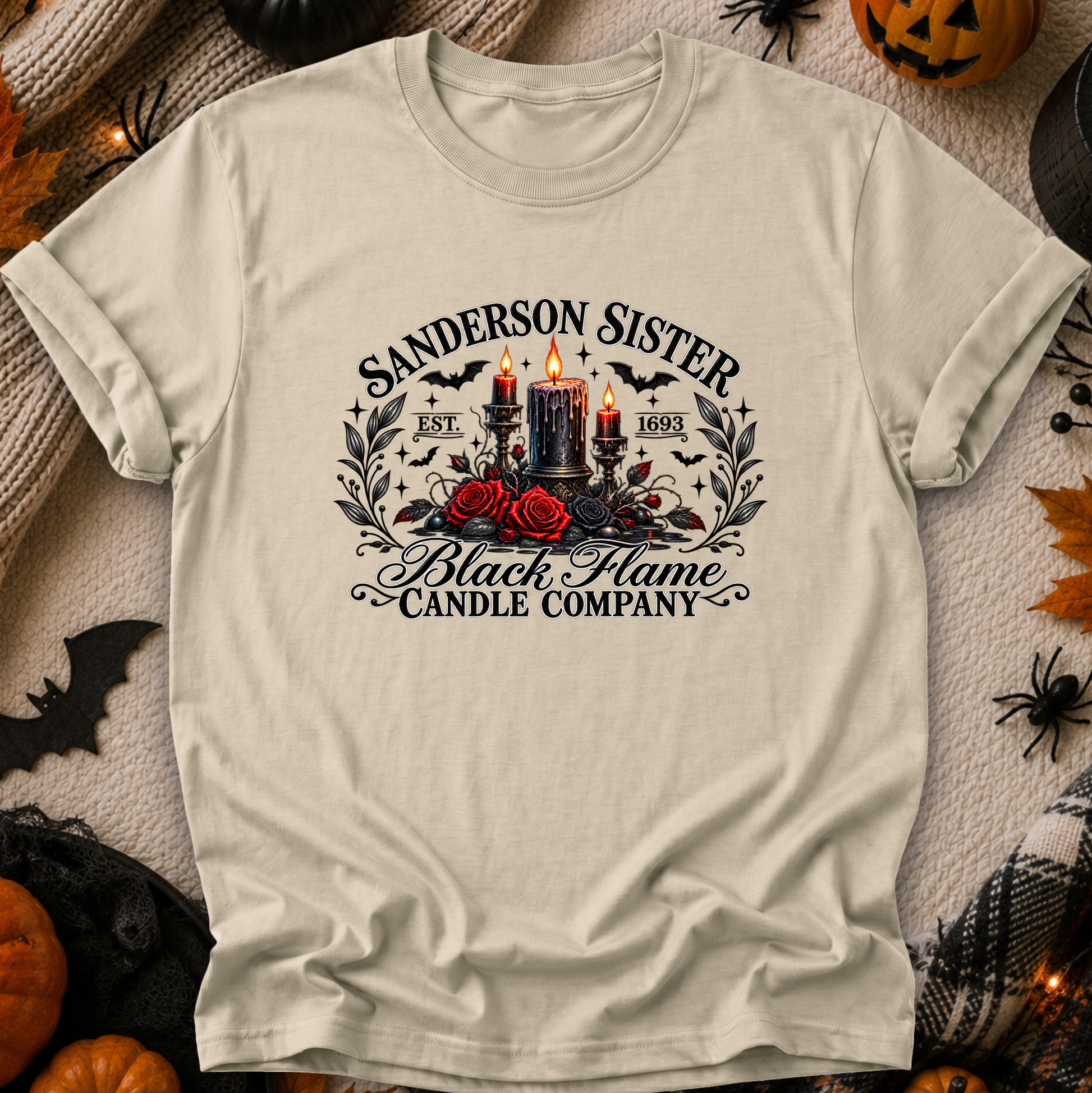 A cream-colored graphic t-shirt laid flat on a textured beige fabric background decorated with Halloween props like pumpkins, spiderwebs, and a black plaid blanket. The t-shirt features a gothic-style graphic with the text 'SANDERSON SISTER' and 'Black Flame Candle Company' surrounding an illustration of two lit black candles, red roses, and bats.