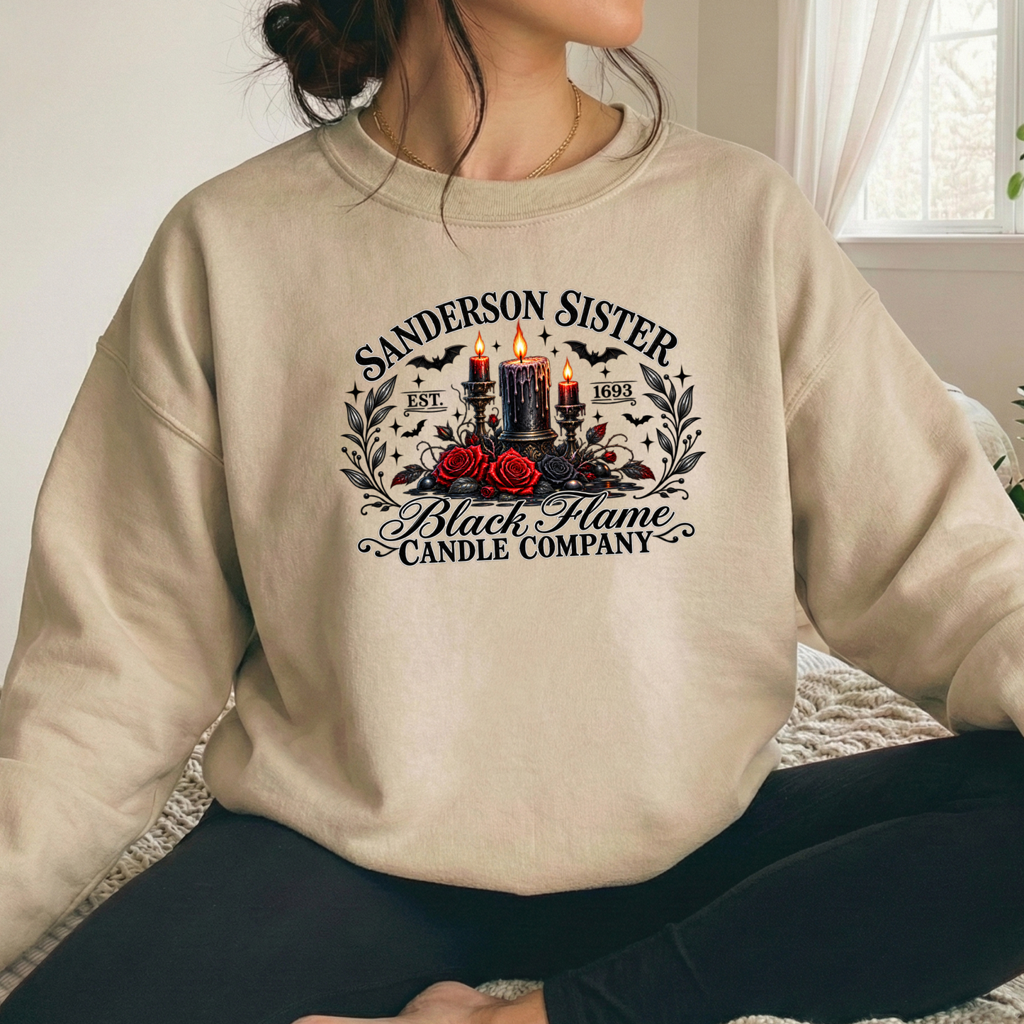 A person wearing a beige crewneck sweatshirt with a gothic-style graphic that reads 'Sanderson Sister Black Flame Candle Company' featuring candles, roses, and bats, paired with black pants in a bright indoor setting.