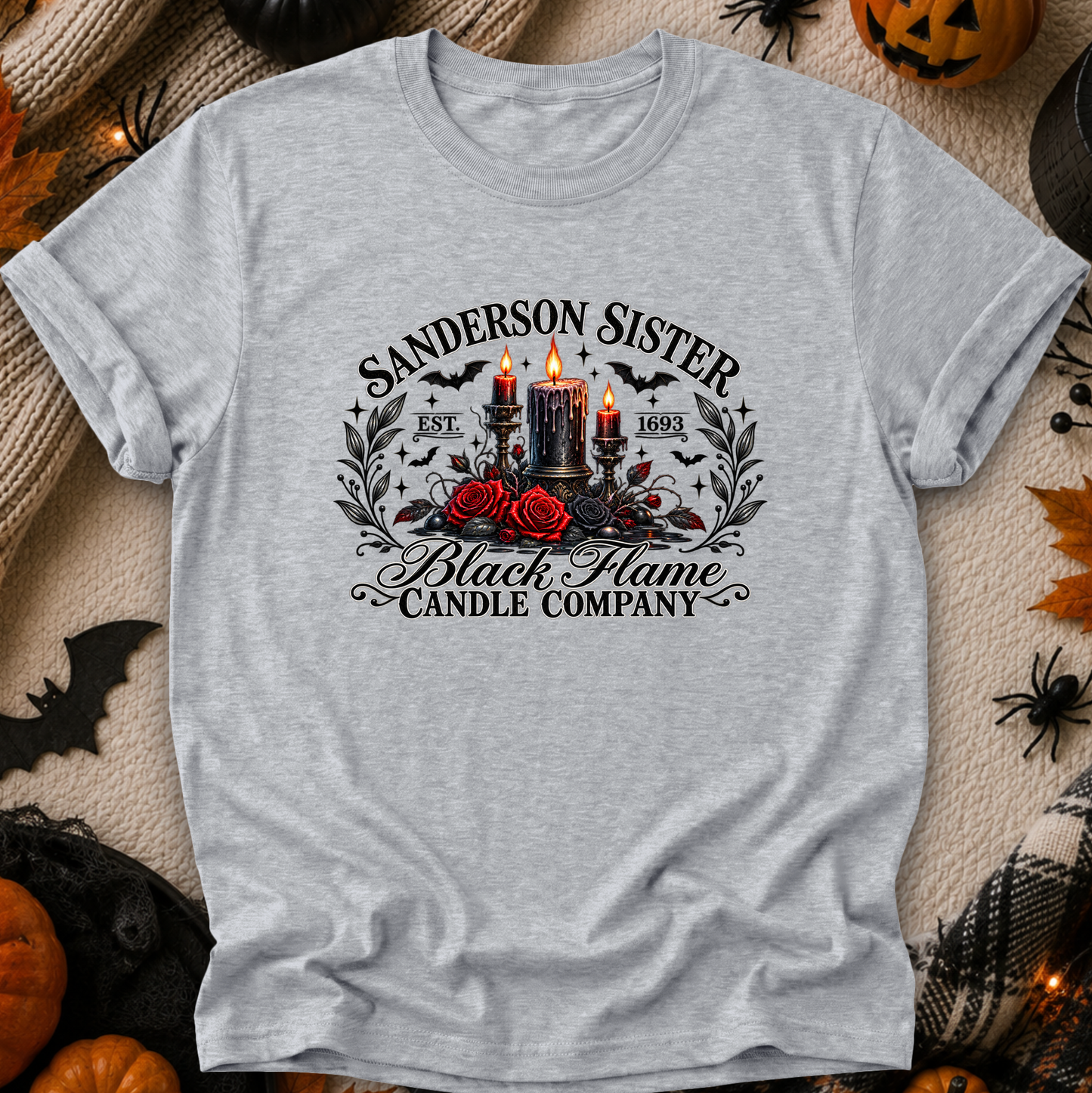 A light heather gray short-sleeve t-shirt with a gothic-themed graphic print reading 'Sanderson Sister Black Flame Candle Company' around an illustration of two lit black candles, red roses, bats, and skulls, laid flat on a cozy autumn-themed background with pumpkins, spider decorations, and leaves.