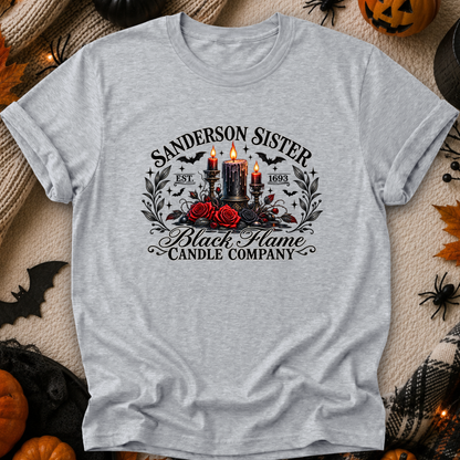 A light heather gray short-sleeve t-shirt with a gothic-themed graphic print reading 'Sanderson Sister Black Flame Candle Company' around an illustration of two lit black candles, red roses, bats, and skulls, laid flat on a cozy autumn-themed background with pumpkins, spider decorations, and leaves.