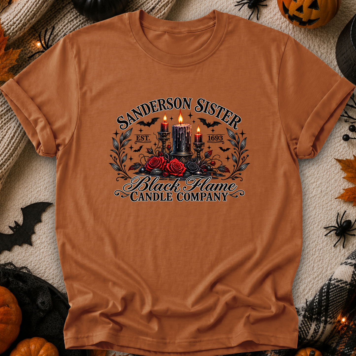A rust-orange graphic t-shirt laid flat on a cozy Halloween-themed background. The shirt features a gothic-style graphic with the text 'SANDERSON SISTER' and 'Black Flame Candle Company' surrounding an illustration of two black candles with bright red flames, red roses, and bats.