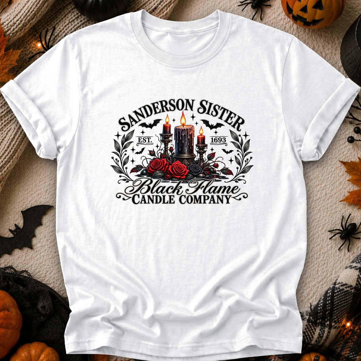 A white short-sleeve graphic T-shirt laid flat on a Halloween-themed background with pumpkins, spider decorations, and black fabric. The shirt features a gothic-style front print reading 'Sanderson Sister Black Flame Candle Company' with black candles, red roses, bats, and the years 1871 and 1895.