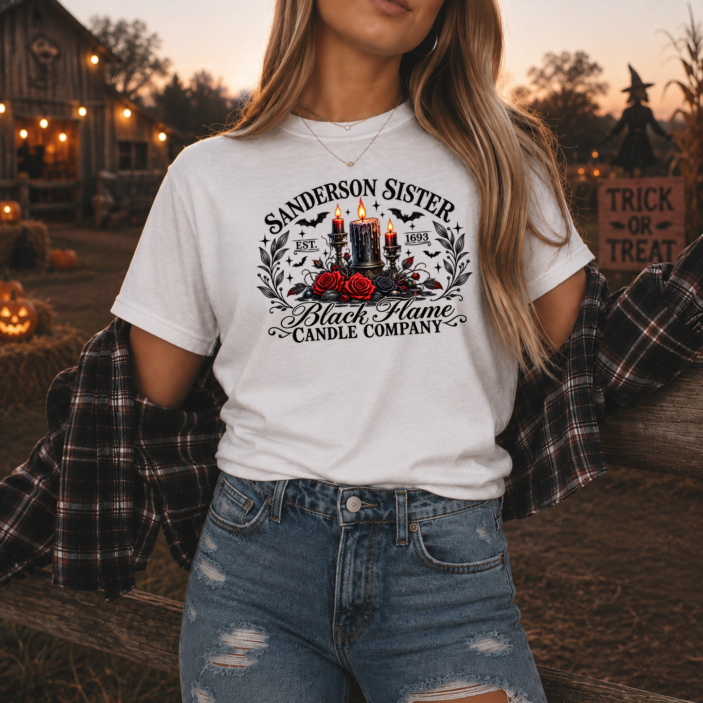 A woman wearing a white graphic t-shirt with a 'Sanderson Sister Black Flame Candle Company' design, styled with a plaid flannel shirt off her shoulders and distressed blue jeans, set against a warm, Halloween-themed outdoor background.