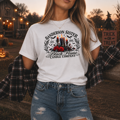 A woman wearing a white graphic t-shirt with a 'Sanderson Sister Black Flame Candle Company' design, styled with a plaid flannel shirt off her shoulders and distressed blue jeans, set against a warm, Halloween-themed outdoor background.