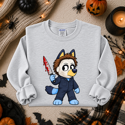 A light gray long-sleeve sweatshirt with a cartoon bluey character graphic holding a bloody knife, styled in a flat lay with Halloween props including pumpkins, spider decorations, a candle, and autumn leaves. The Gildan brand label is visible inside the collar.