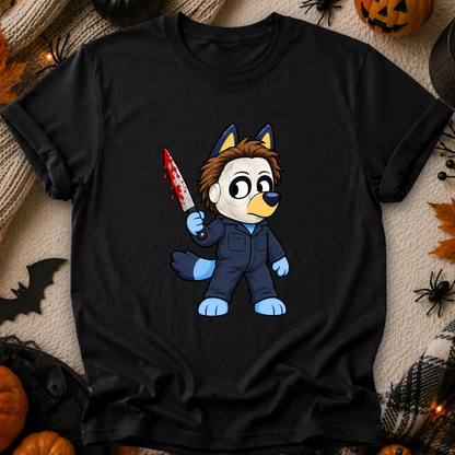 A black short-sleeve graphic T-shirt laid flat on a cozy beige fabric background with Halloween props including pumpkins, spiders, and a bat. The shirt features a cartoon wolf character in a blue jumpsuit holding a blood-splattered knife.