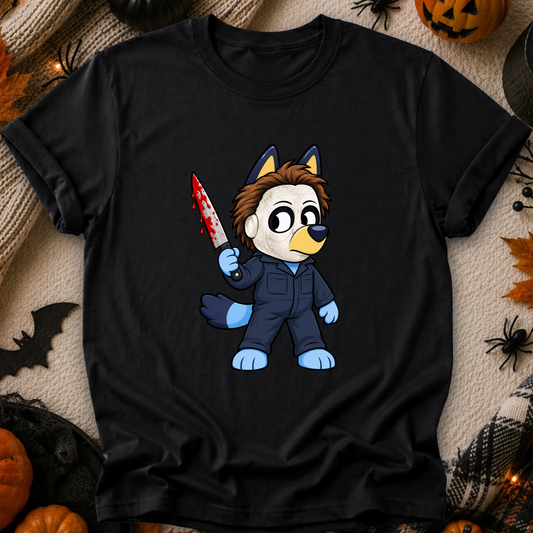 A black short-sleeve graphic T-shirt laid flat on a cozy beige fabric background with Halloween props including pumpkins, spiders, and a bat. The shirt features a cartoon wolf character in a blue jumpsuit holding a blood-splattered knife.