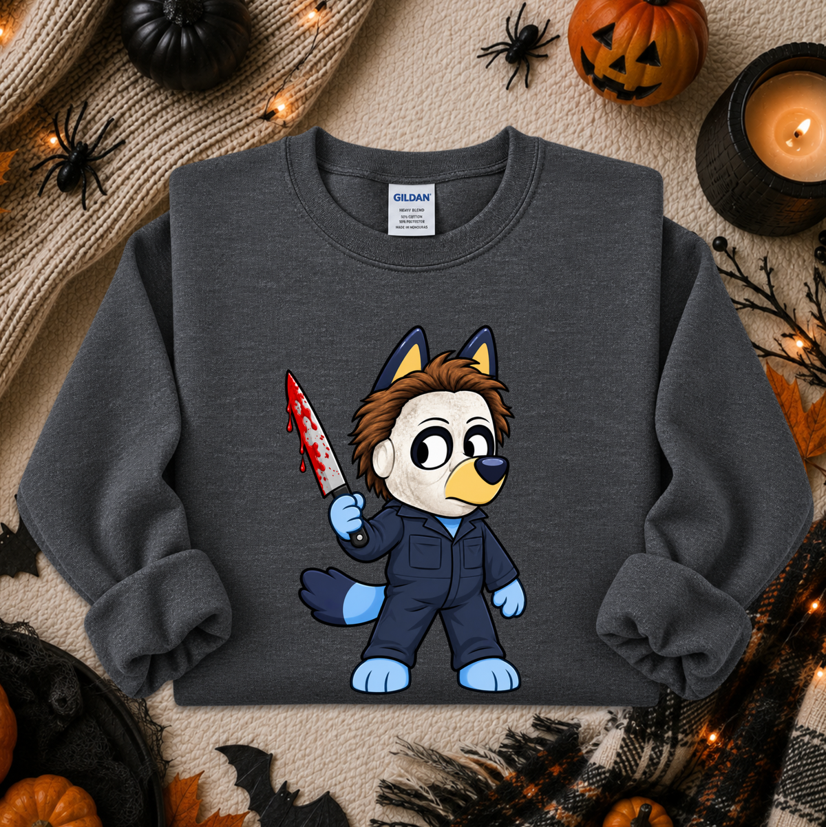 A dark gray crewneck sweatshirt featuring a cartoon graphic of a dog character in a blue jumpsuit holding a bloody knife. The sweatshirt is displayed in a flat-lay Halloween setting with pumpkins, a candle, and spider decorations.