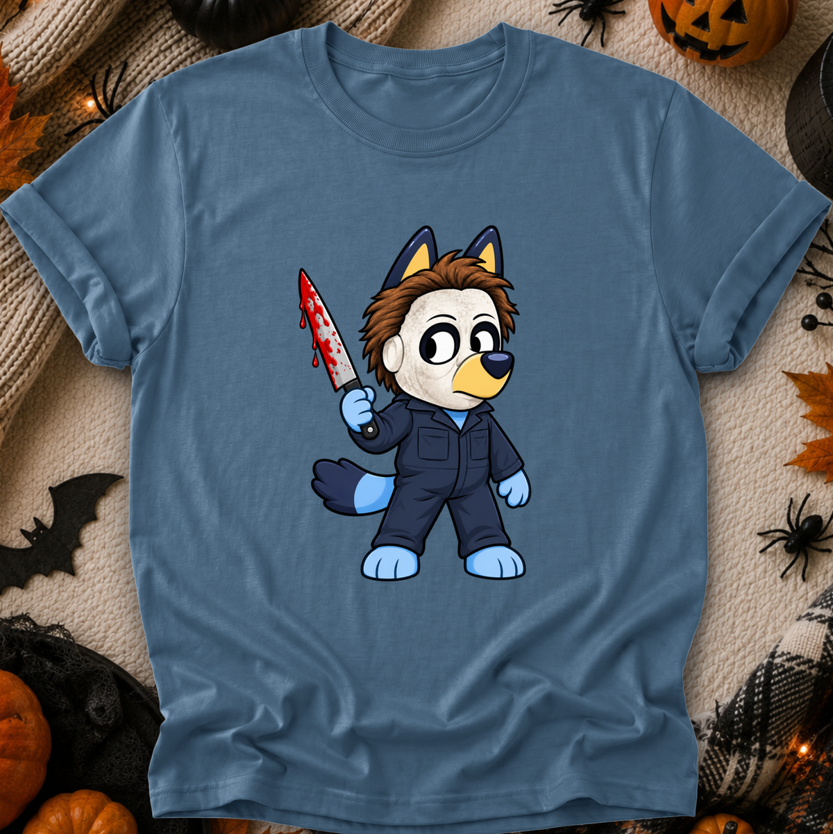 A blue crewneck t-shirt featuring a cartoon graphic of a bluey character in a dark jumpsuit holding a bloody knife, styled in a flat lay with Halloween decorations like pumpkins, a spider, and a black spiderweb on a beige fabric background.