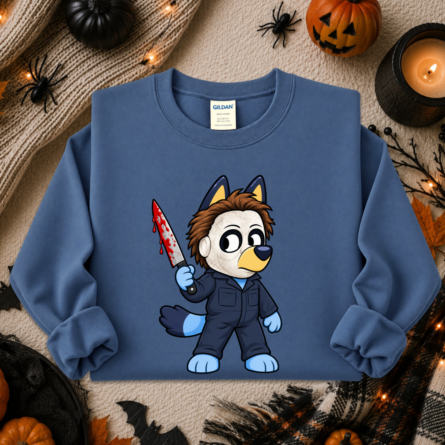 A blue crewneck sweatshirt featuring a cartoon graphic of a dog character in a black jumpsuit holding a bloody knife, styled in a cozy Halloween flat-lay with pumpkins, a candle, and spider decorations.