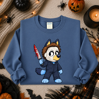 A blue crewneck sweatshirt featuring a cartoon graphic of a dog character in a black jumpsuit holding a bloody knife, styled in a cozy Halloween flat-lay with pumpkins, a candle, and spider decorations.