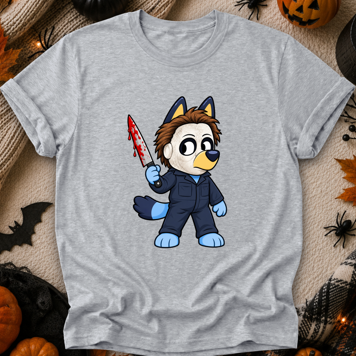 A light heather gray short-sleeve t-shirt with a cartoon graphic of a bluey character in a blue jumpsuit holding a blood-splattered knife, styled in a flat lay with Halloween props like pumpkins, a spider, and a black spiderweb.