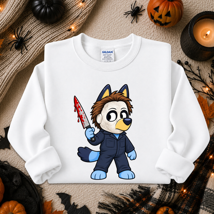 A white long-sleeve crewneck sweatshirt featuring a cartoon graphic of a dog character in a blue jumpsuit holding a bloody knife. The sweatshirt is displayed in a flat-lay Halloween setting with pumpkins, a candle, spider decorations, and a cozy knit blanket.