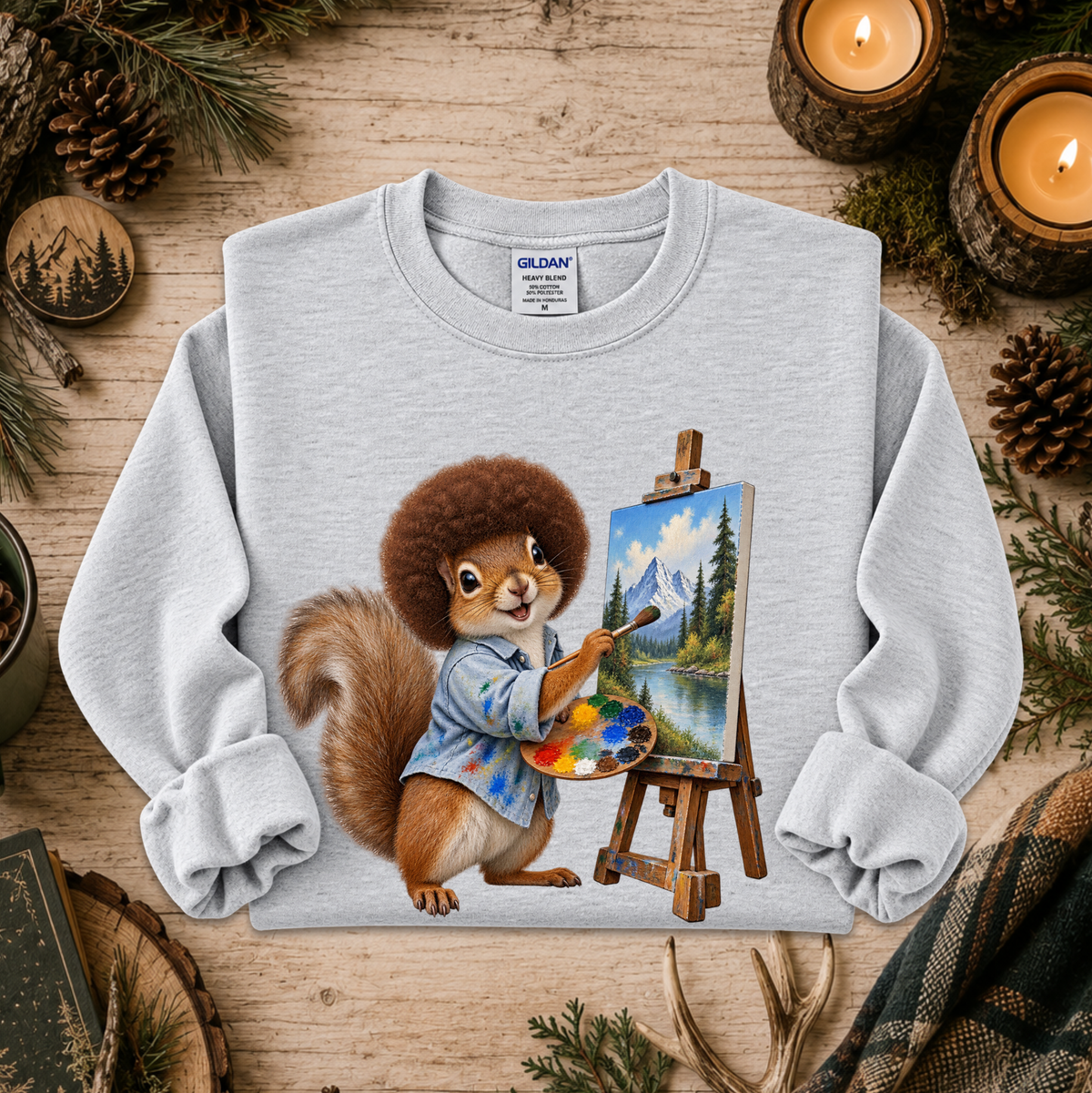 A light gray crewneck sweatshirt featuring a colorful graphic of a squirrel dressed as an artist painting a mountain landscape on an easel. The sweatshirt is displayed in a flat-lay setting on a rustic wooden surface surrounded by pinecones, evergreen branches, and burning candles.