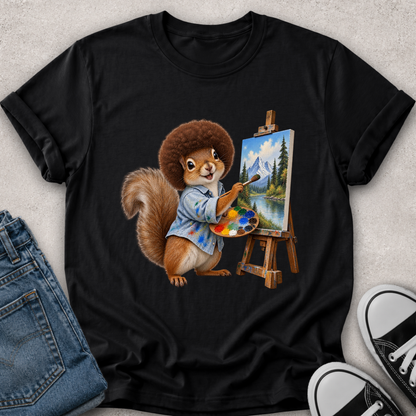 A black short-sleeve graphic T-shirt laid flat on a light textured surface, styled with blue jeans on the left and black-and-white sneakers on the right. The shirt features a colorful illustration of a squirrel painting a mountain landscape on an easel.