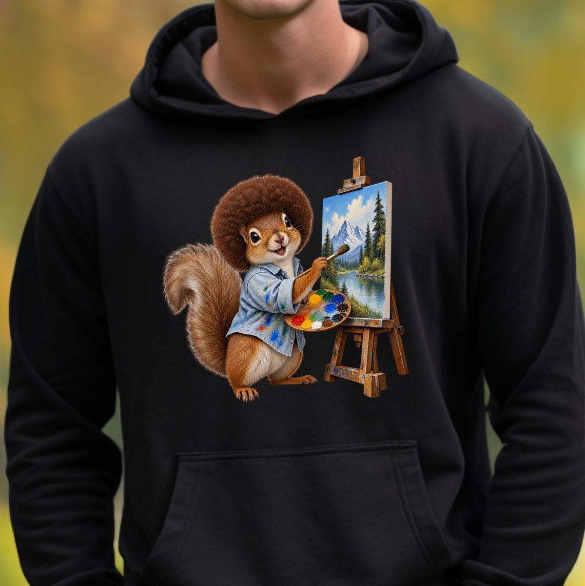 A person wearing a black pullover hoodie with a colorful graphic of a squirrel painting at an easel on the front, set against a blurred outdoor background.