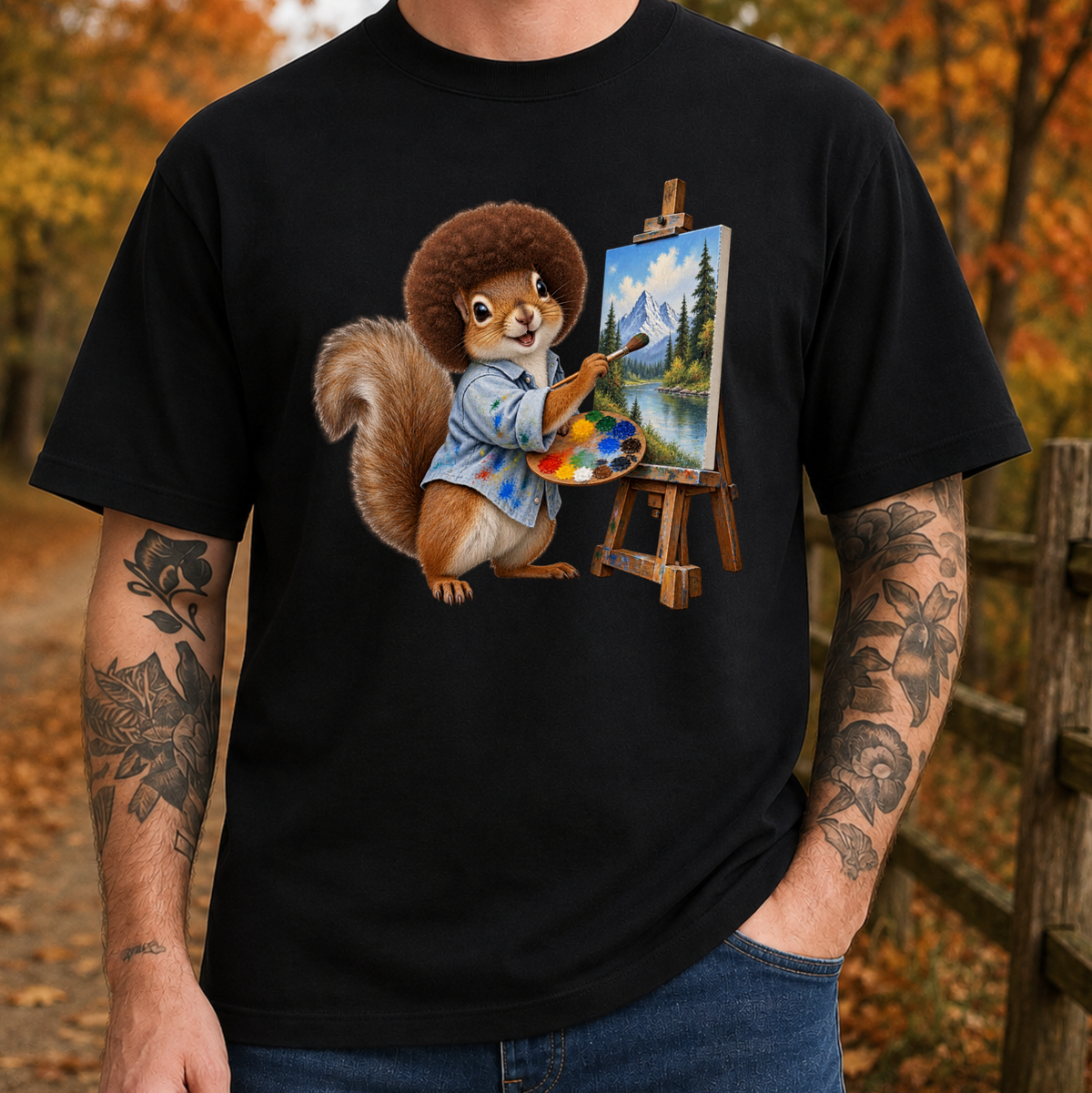 A man wearing a black crewneck t-shirt with a colorful graphic of a squirrel painting a landscape on an easel, set against an autumn outdoor background.