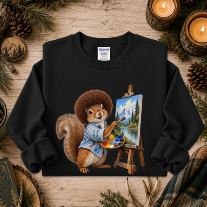 A black crewneck sweatshirt featuring a colorful graphic of a squirrel painting a mountain landscape on an easel, styled on a rustic wooden background with pinecones, candles, and evergreen branches.