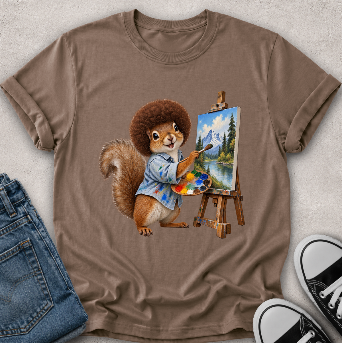 A brown short-sleeve crewneck t-shirt laid flat on a light gray textured surface, featuring a graphic of a squirrel painting a mountain landscape on an easel. Blue jeans and black-and-white sneakers are styled around the shirt.