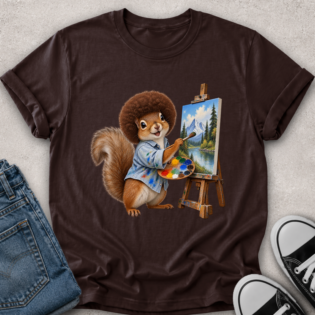 A dark brown short-sleeve t-shirt laid flat on a light textured surface, featuring a colorful graphic of a squirrel painting a mountain landscape on an easel. Blue jeans and black-and-white sneakers are partially visible as styling props.