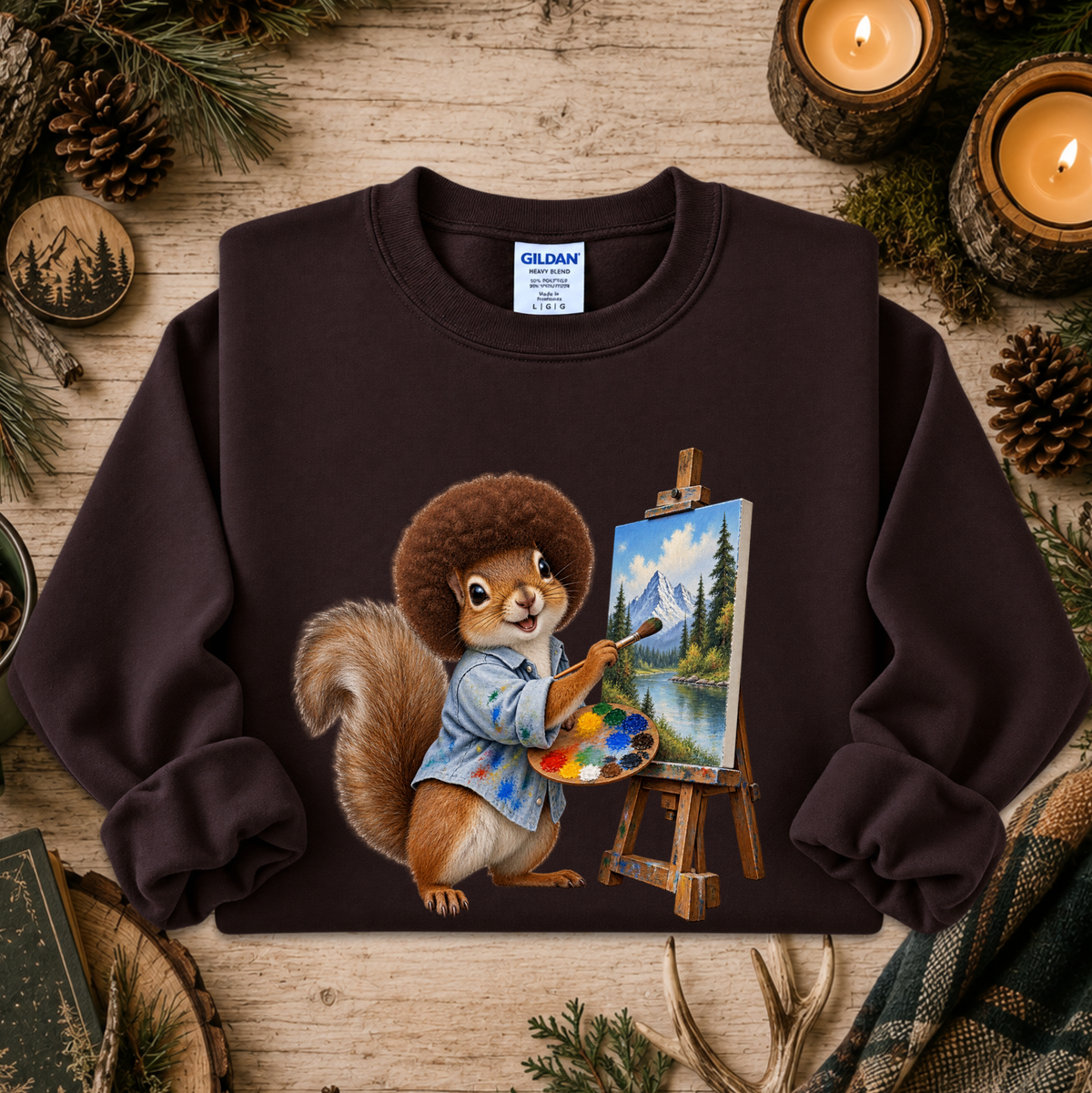 A dark brown crewneck sweatshirt featuring a graphic of a squirrel painting a landscape on an easel, styled on a rustic wooden background with pine needles, candles, and pinecones.