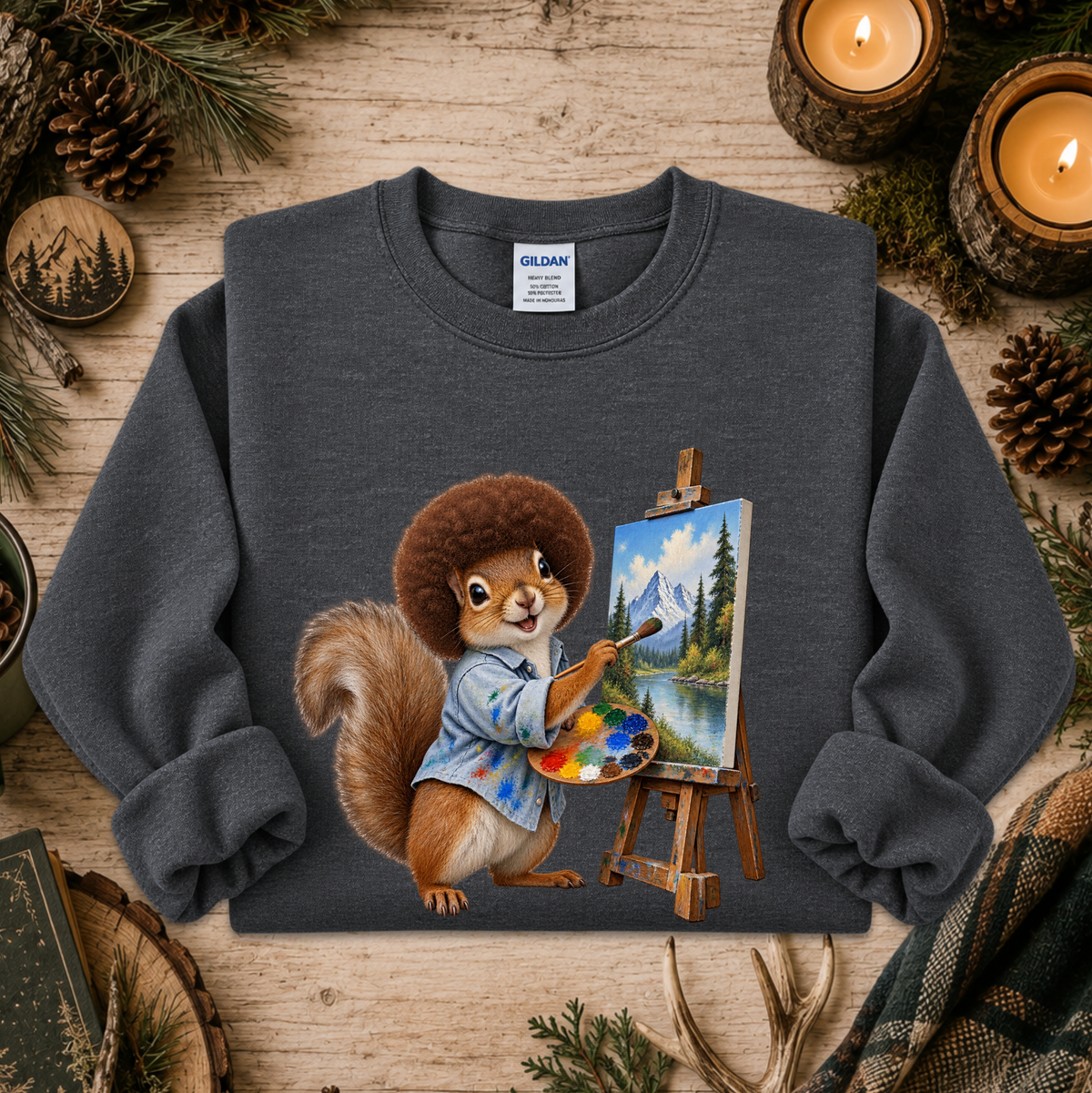 A dark gray crewneck sweatshirt featuring a graphic of a cartoon squirrel painting a mountain landscape on an easel. The sweatshirt is displayed in a flat-lay setting on a rustic wooden surface, surrounded by pine needles, pinecones, and burning candles.