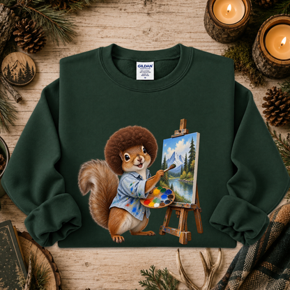 A dark green crewneck sweatshirt featuring a graphic of a squirrel painting a mountain landscape on an easel, styled in a flat-lay on a rustic wooden background with pinecones, candles, and evergreen branches.