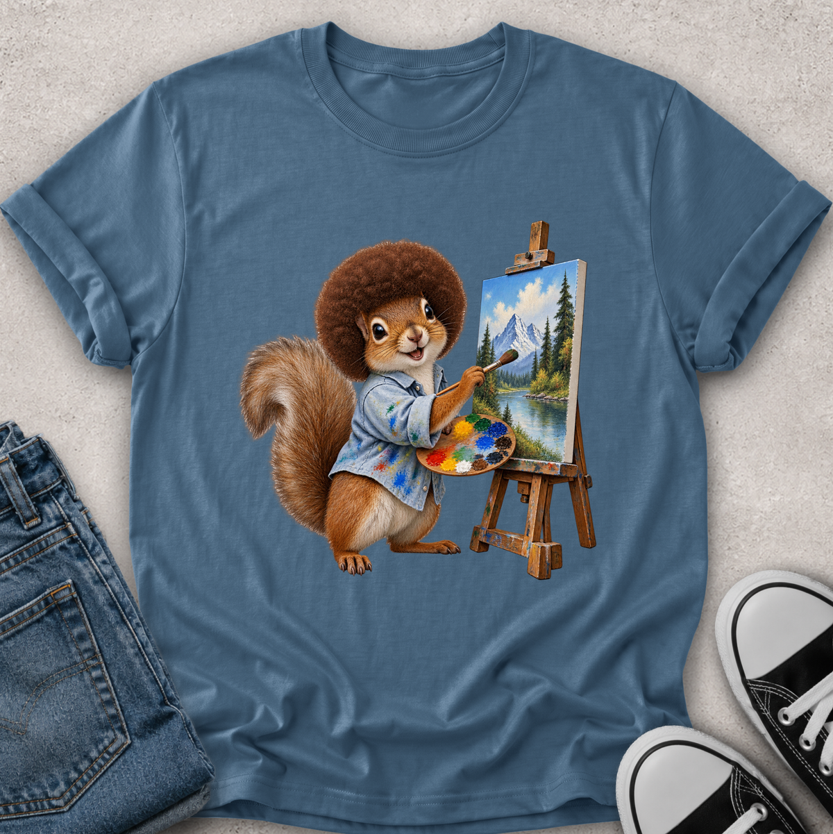A blue short-sleeve crewneck t-shirt laid flat on a light textured surface, featuring a graphic of a squirrel painting a mountain landscape on an easel. The shirt is styled with blue jeans on the left and black-and-white sneakers on the right.