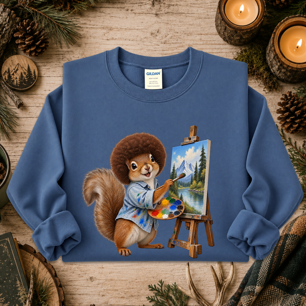 A blue crewneck sweatshirt featuring a printed graphic of a squirrel painting a mountain landscape on an easel, styled on a rustic wooden background with pine branches, candles, and a plaid scarf.