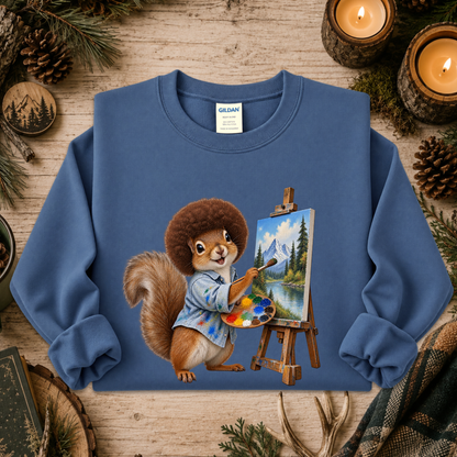 A blue crewneck sweatshirt featuring a printed graphic of a squirrel painting a mountain landscape on an easel, styled on a rustic wooden background with pine branches, candles, and a plaid scarf.