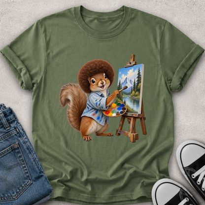 An olive green short-sleeve t-shirt laid flat on a light textured surface, featuring a cartoon squirrel painting a mountain landscape on an easel. Blue jeans and black-and-white sneakers are partially visible as styling props.