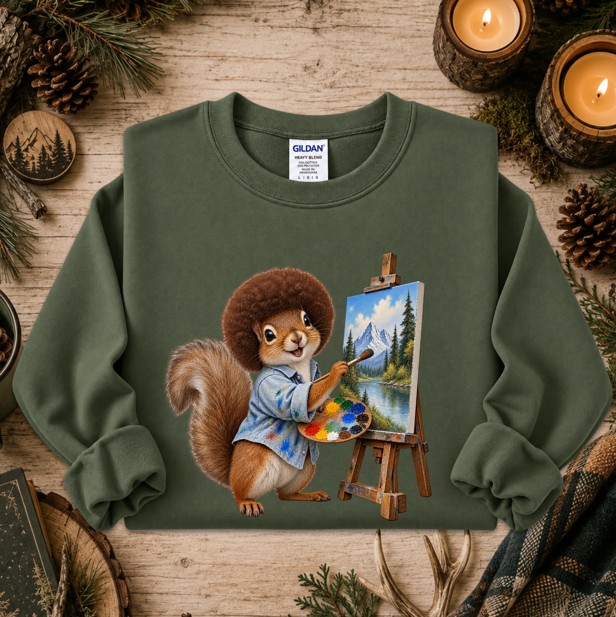 An olive green crewneck sweatshirt by Gildan, featuring a printed graphic of a cartoon squirrel painting a mountain landscape on an easel. The sweatshirt is displayed in a flat-lay setting on a rustic wooden surface, surrounded by pine needles, pinecones, lit candles, and a plaid scarf.