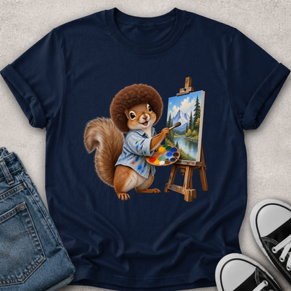 A navy blue short-sleeve t-shirt laid flat on a light textured surface, featuring a colorful graphic of a squirrel painting a mountain landscape on an easel. Folded blue jeans and black-and-white sneakers are partially visible around the shirt as styling props.