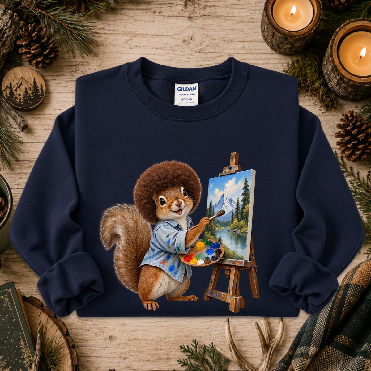 A navy blue crewneck sweatshirt with a graphic of a squirrel painting at an easel, styled on a rustic wooden background with pine branches, candles, and pinecones. The neck label shows Gildan.