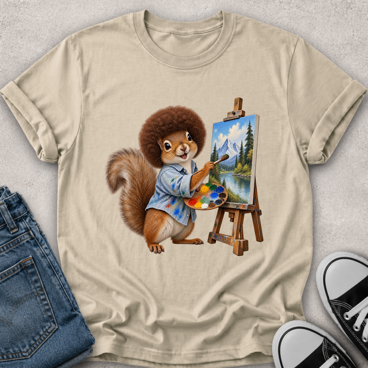 A beige graphic t-shirt featuring a cartoon illustration of a squirrel painting a mountain landscape on an easel, styled in a flat-lay with blue jeans and black sneakers on a light gray textured background.