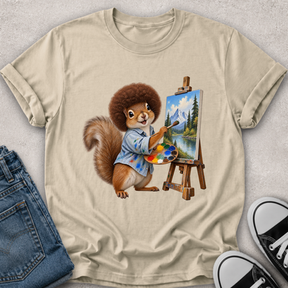A beige graphic t-shirt featuring a cartoon illustration of a squirrel painting a mountain landscape on an easel, styled in a flat-lay with blue jeans and black sneakers on a light gray textured background.