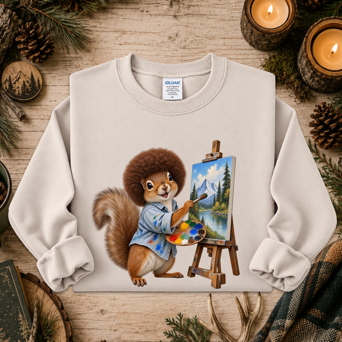 A light beige crewneck sweatshirt featuring a colorful graphic of a squirrel painting in a blue shirt on an easel, laid flat on a rustic wooden surface decorated with pine branches, candles, and pinecones.