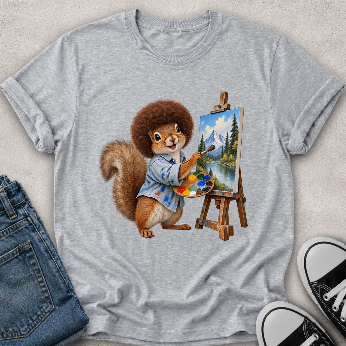 A heather gray short-sleeve t-shirt with a graphic of a cartoon squirrel painting a mountain landscape on an easel, styled in a flat lay with blue jeans and black-and-white sneakers on a light textured background.