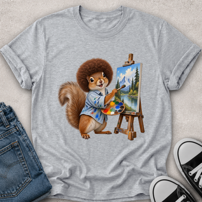 A heather gray short-sleeve t-shirt with a graphic of a cartoon squirrel painting a mountain landscape on an easel, styled in a flat lay with blue jeans and black-and-white sneakers on a light textured background.