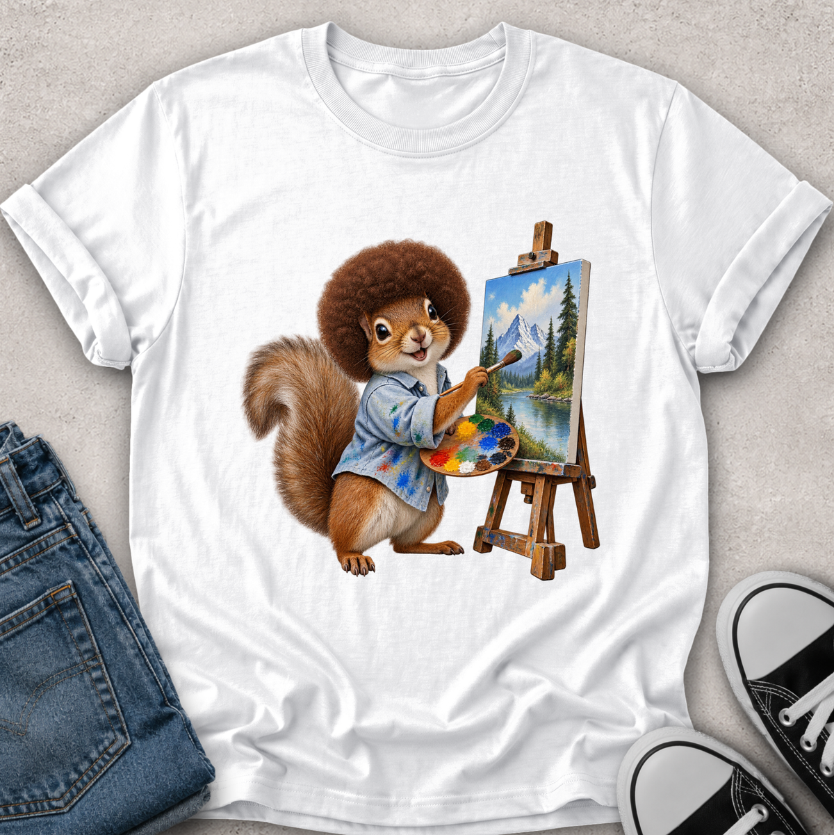 A white short-sleeve crewneck t-shirt laid flat on a light gray textured surface, featuring a colorful graphic of a cartoon squirrel painting a mountain landscape on an easel. Folded blue jeans and black-and-white sneakers are styled around the shirt.