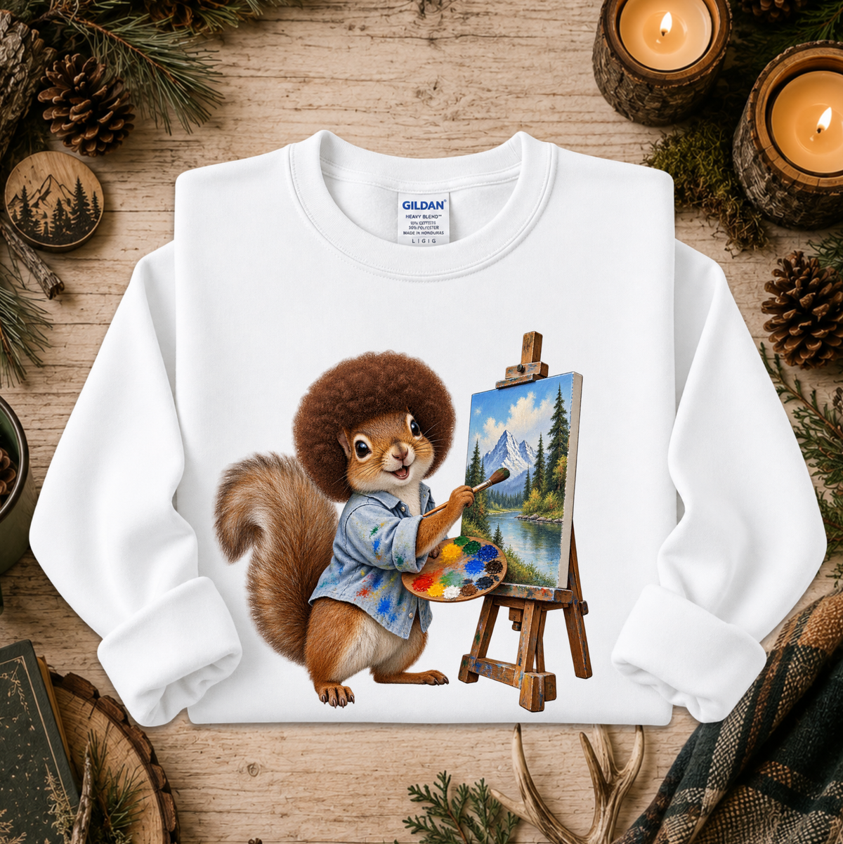 A white long-sleeve crewneck sweatshirt laid flat on a rustic wooden surface, featuring a colorful graphic of a squirrel painting a mountain landscape on an easel. The sweatshirt is styled with pine branches, candles, and pinecones.