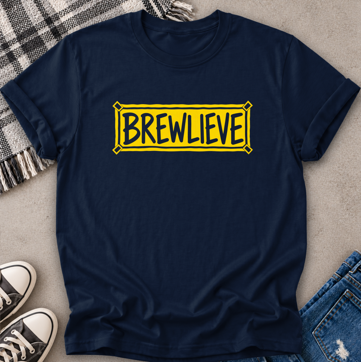 A navy blue crewneck t-shirt with a yellow 'BREWLIEVE' graphic printed on the chest, styled in a flat-lay arrangement with jeans, sneakers, and a plaid blanket on a concrete floor.