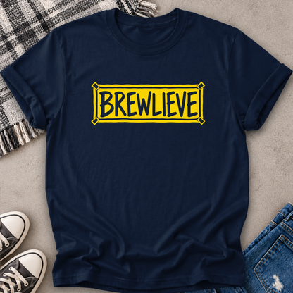 A navy blue crewneck t-shirt with a yellow 'BREWLIEVE' graphic printed on the chest, styled in a flat-lay arrangement with jeans, sneakers, and a plaid blanket on a concrete floor.