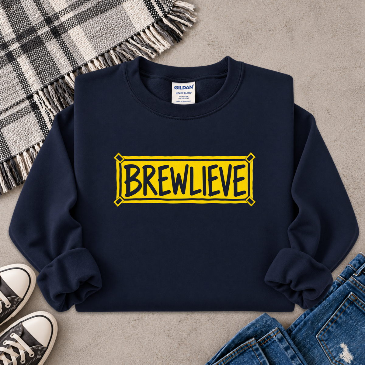 A navy blue crewneck sweatshirt with a yellow sign graphic that reads 'BREWLIEVE' in black letters, styled in a flat lay with jeans, a plaid blanket, and sneakers on a concrete floor.