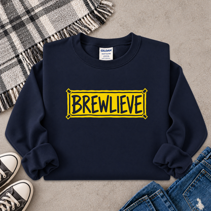 A navy blue crewneck sweatshirt with a yellow sign graphic that reads 'BREWLIEVE' in black letters, styled in a flat lay with jeans, a plaid blanket, and sneakers on a concrete floor.