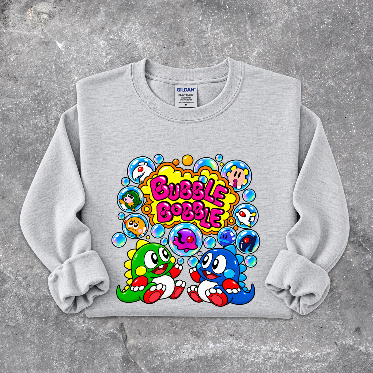 A light gray crewneck sweatshirt with a colorful graphic print featuring cartoon characters and the text 'Bubble Bubble'. The sweatshirt is laid flat on a textured gray concrete surface.
