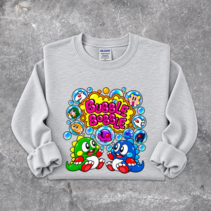 A light gray crewneck sweatshirt with a colorful graphic print featuring cartoon characters and the text 'Bubble Bubble'. The sweatshirt is laid flat on a textured gray concrete surface.