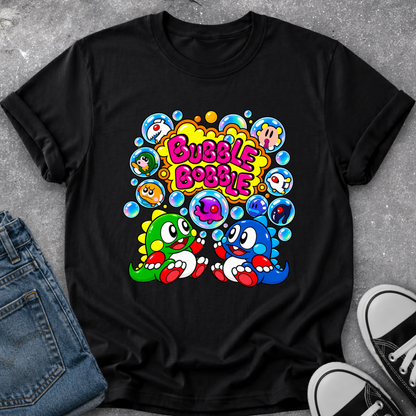 A black short-sleeve graphic T-shirt laid flat on a gray textured surface, styled with folded blue jeans on the left and black-and-white sneakers on the right. The shirt features a colorful retro-style bubble graphic with the words “Bubble Bobble” in pink and yellow, along with cartoon characters and blue bubbles.